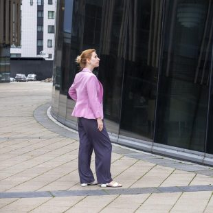 Young blonde woman with tailored hairstyle in pink jacket stands in front of modern office building made of glass and steel and looks at her reflection. Profile view. Selective focus.